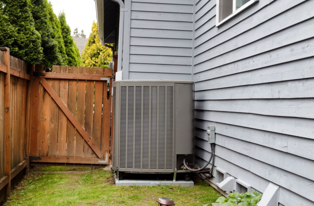 Outdoor air conditioning unit next to a house with gray siding and a wooden fence.