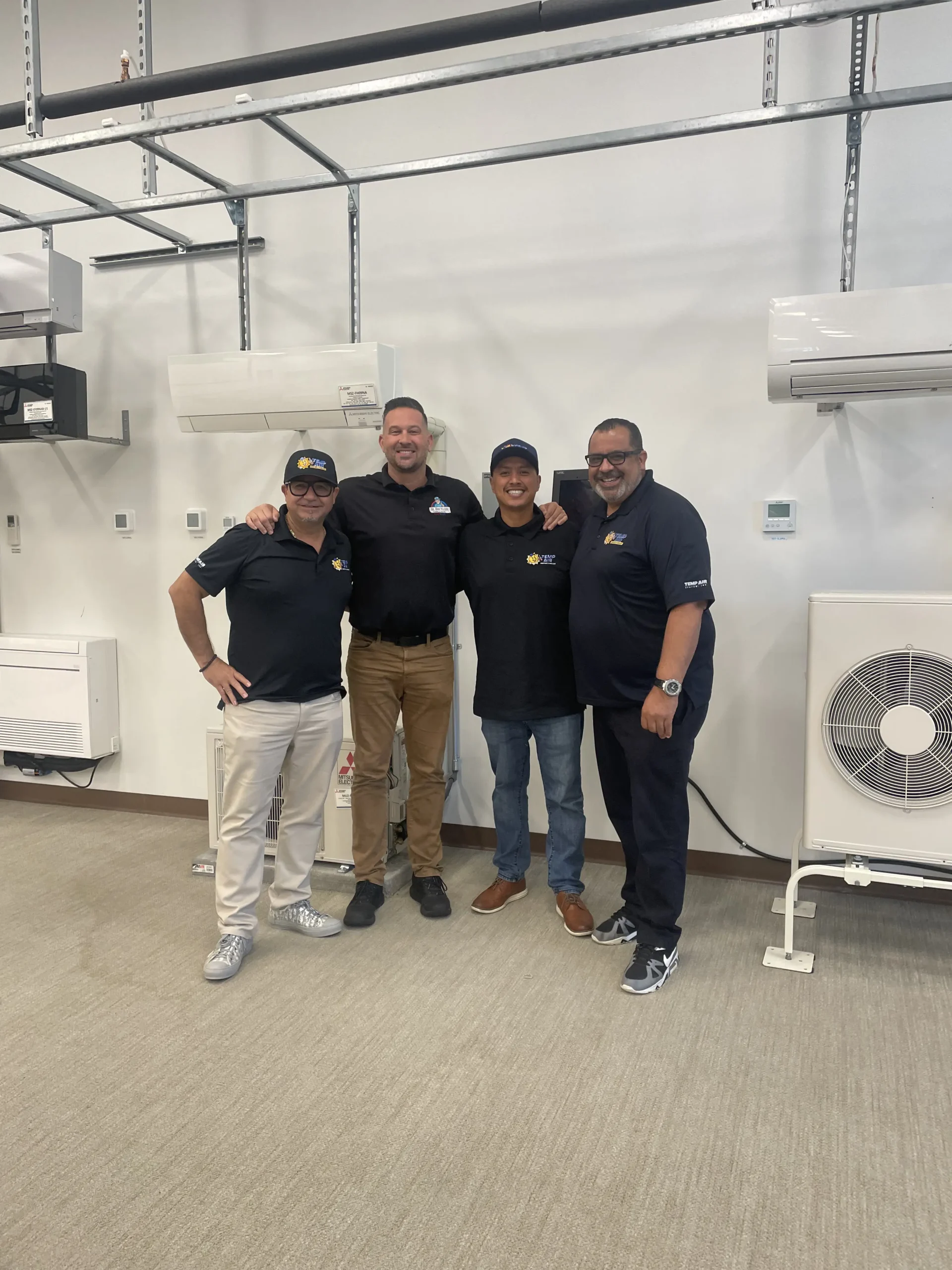 Four technicians in front of various air conditioning units and HVAC equipment.