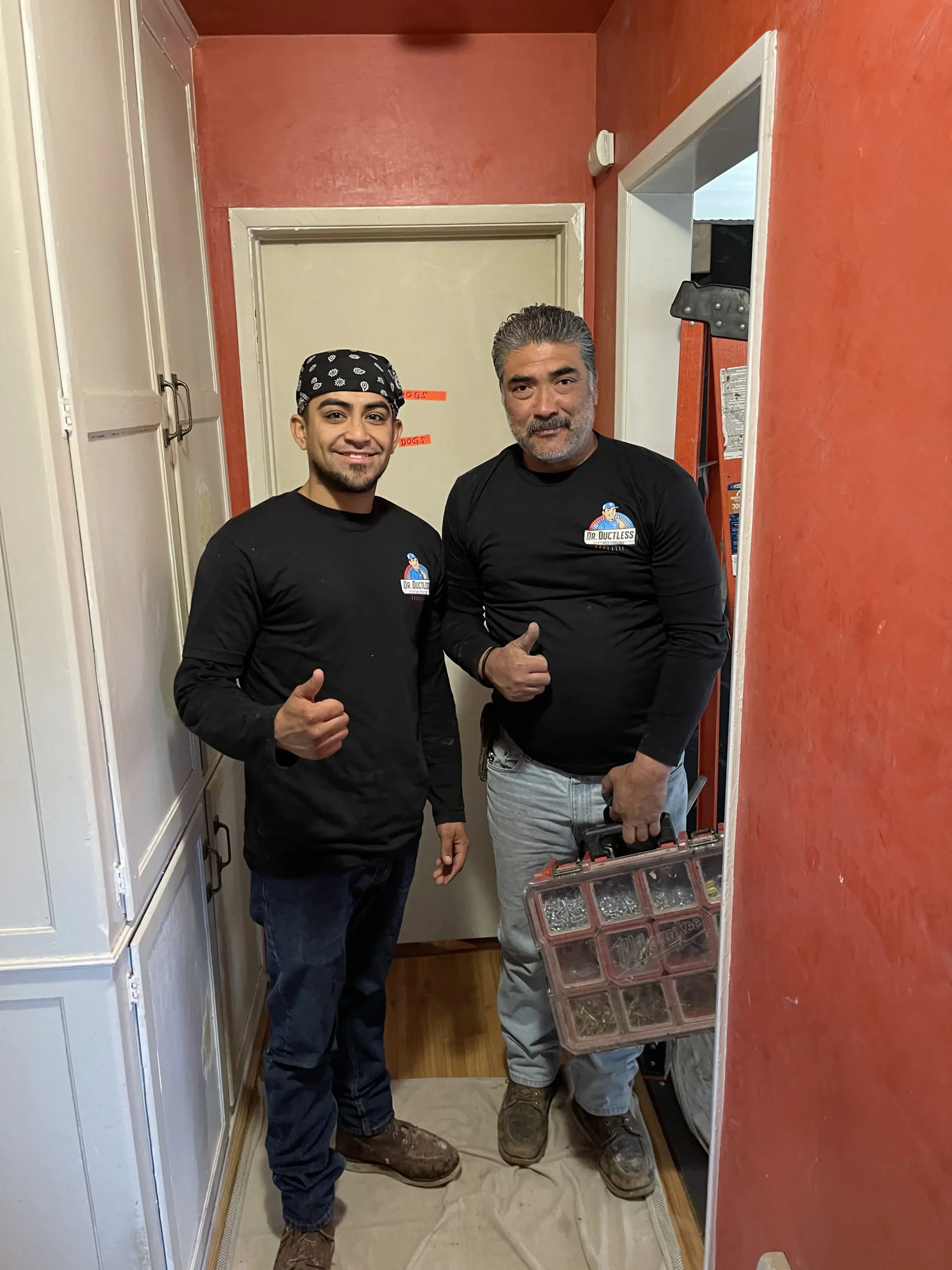 Two workers in black shirts with company logos give thumbs up in a narrow hallway.